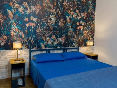 Camera matrimoniale con letto blu, comodini con lampade e parete in mattoncini chiari sotto la carta da parati floreale